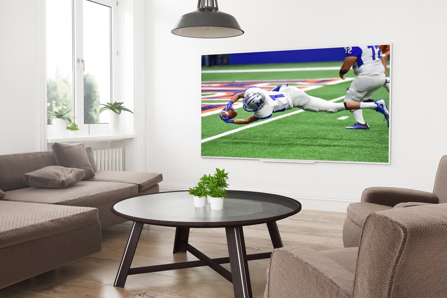 A modern living room with a large wall-mounted TV displaying a football game, featuring comfortable seating, neutral tones, and natural light from large windows.